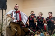 Mężczyzna grający na akordeonie, obok jednakowo ubrane panie z zespołu wokalnego 